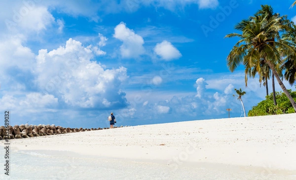 Fototapeta Back view of man in straw hat walking alone on white sandy beach on maldives island with blue sky at background. Solo travel, vacation, wide angle