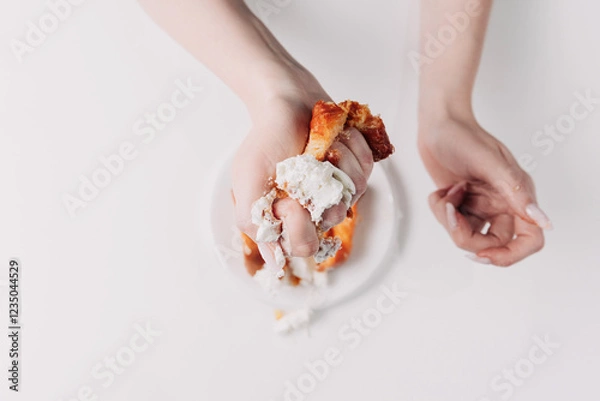 Fototapeta Woman's hands tearing, unwrapping, and crushing a pastry on a white plate against a white background