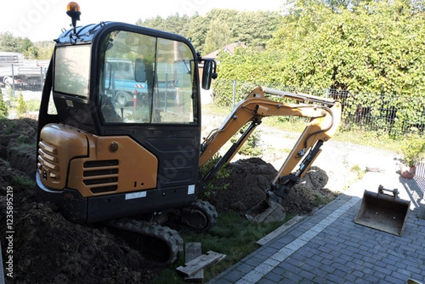 Fototapeta A black and yellow compact hydraulic excavator standing in a courtyard next to heap of dirt