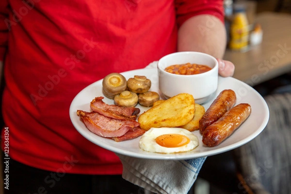 Obraz Full Irish breakfast served by waiter at a hotel with sausge, eggs, baked beans, bacon, hash browns and mushrooms.