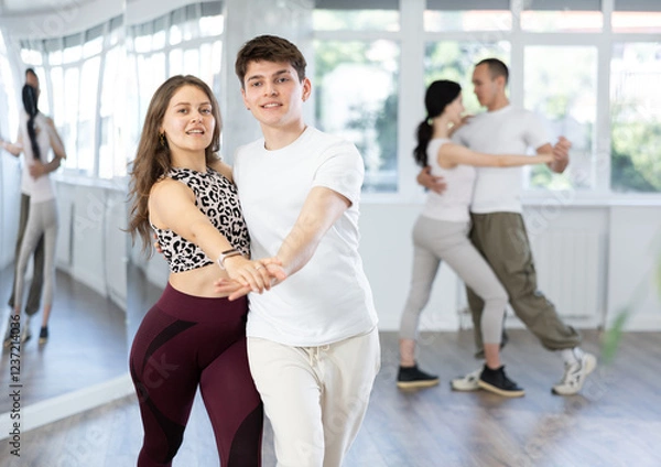 Fototapeta Couple of smiling young guy and girl enjoying slow foxtrot in dance studio. Amateur ballroom dancing concept..