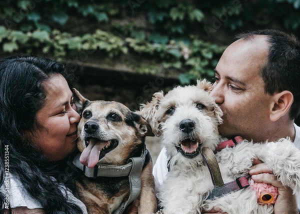 Fototapeta A smiling couple hugs and kisses their two dogs, who look happy with their tongues out. The background has green leaves, creating a warm and loving atmosphere.
