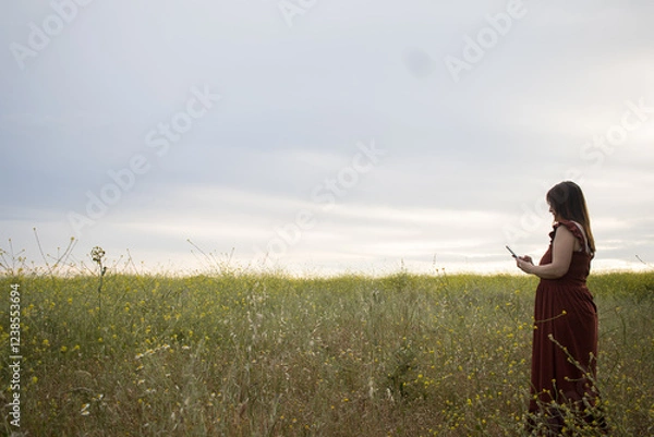 Obraz Shot of a woman using a cellphone in a field. Lady holding a smartphone looking sideways. Horizontal
