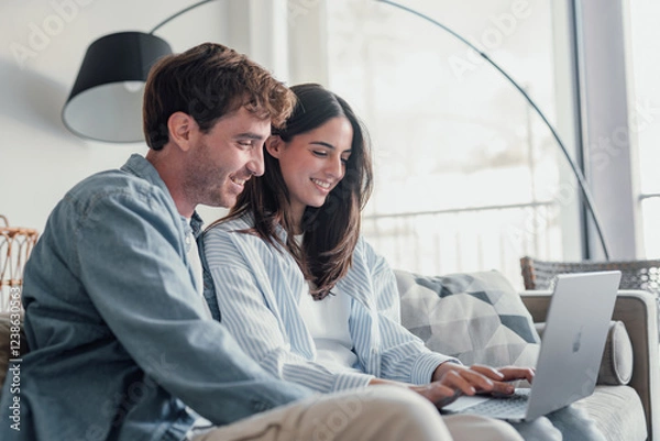 Fototapeta Happy millennial couple sit relax on couch in living room watching video on laptop together, smiling young husband and wife rest on sofa at home browsing Internet using modern computer device.