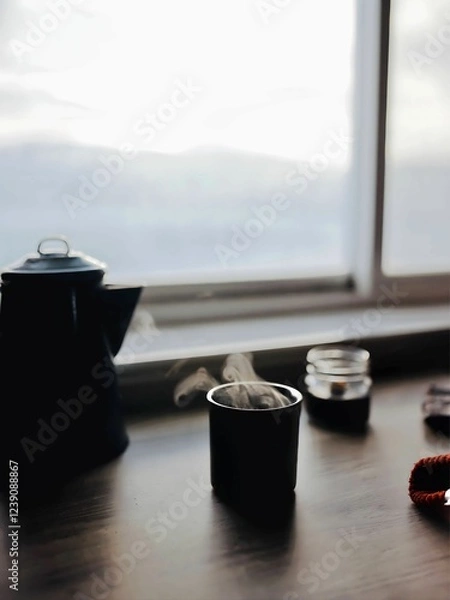 Fototapeta A cozy winter scene with steaming tea, a lit candle, and a knitted scarf resting on a wooden table by a window, creating a warm and inviting atmosphere