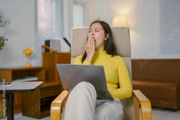 Fototapeta Tired freelancer yawning while working from home sitting on armchair with laptop on legs