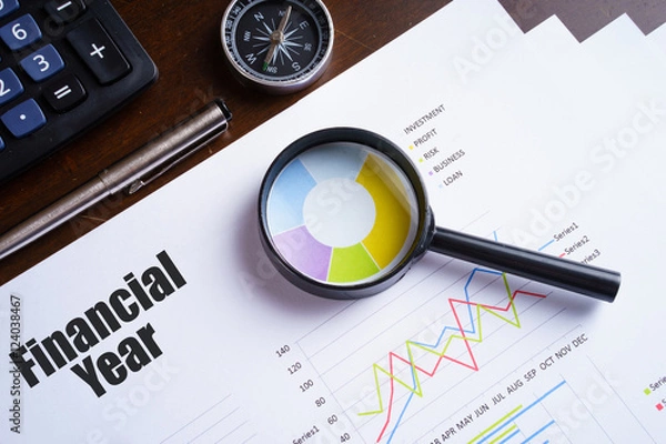 Fototapeta Tekst „Rok finansowy” na arkuszu papieru z lupą na wykresie, kostki do gry, okulary, długopis, laptop i niebiesko-żółta pinezka na drewnianym stole - koncepcja biznesowa, bankowość, finanse i inwestycje