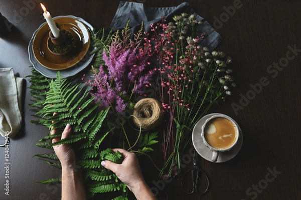 Fototapeta top view of florist woman hands making bouquet of summer garden flowers and ferns on rustic wooden table