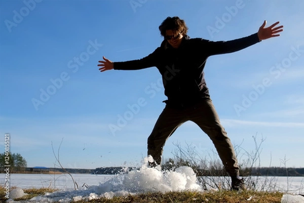 Fototapeta Playful man breaks a block of ice near frozen river on nature in sunny day. Guy throwing ice on the land to break it up. Large piece of ice breaks up into small fragments. Winter, early spring season.