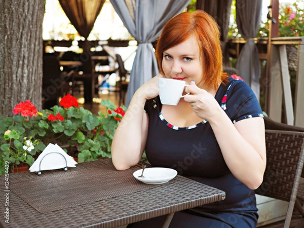Fototapeta Portrait of attractive plus size redhead woman sitting in a cafe in city park with cup of coffee