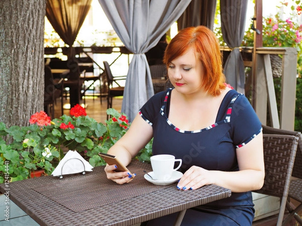 Fototapeta Portrait of attractive plus size redhead woman sitting in a cafe in city park with cup of coffee and checks her smart phone