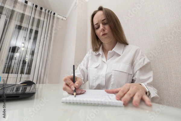 Fototapeta Female person office worker or freelancer works in office or at home, concentrated draws graph or writes something in notebook, work computer is on table next to she. Business woman while working.