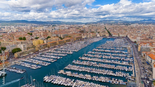 Fototapeta Aerial view of Marseille, France, showcasing the Old Port filled with yachts, surrounded by historic buildings, and a sprawling cityscape extending to the mountains under a partly cloudy blue sky