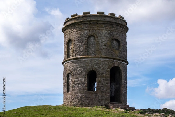 Obraz Grin Low Buxton , England - APRIL 18, 2022: with a pointed roof sits on a hill. The building is empty and has no windows. The sky is cloudy and the grass is green