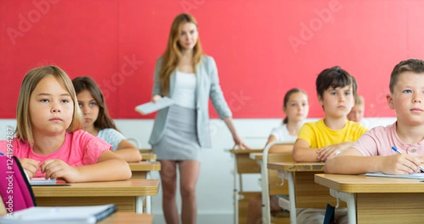 Fototapeta Schoolkids sitting at desks in classroom. They're listening to feamle teacher who's explaining them lesson information.