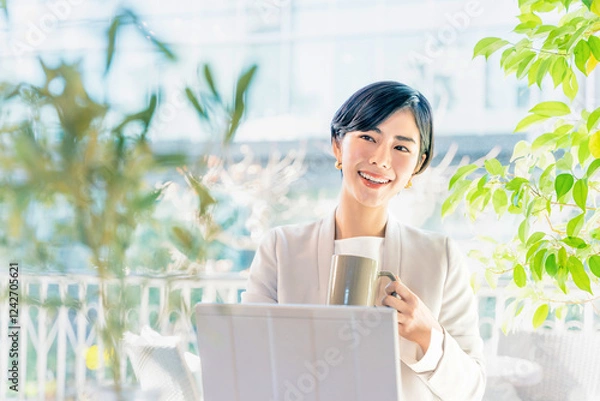 Obraz 明るいオフィスでパソコンの作業中にカップの飲み物を飲んでリラックスする笑顔の女性