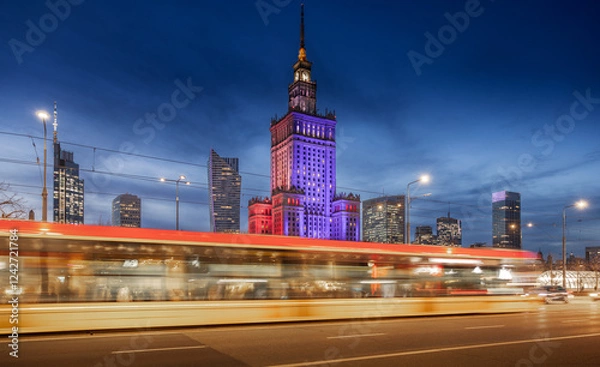 Fototapeta Warsaw Palace of Culture glows in red, white, and blue against the night sky, surrounded by modern skyscrapers. Light trails from passing cars and trams create a dynamic sense of motion 