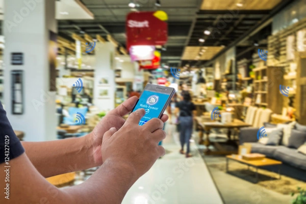 Obraz Płatność mobilna, koncepcja społeczeństwa bezgotówkowego. Ręcznie trzymanie smartfona z płatnością mobilną na ekranie i NFC sygnałów ikon przeciwko abstrakcyjnym meble MART.
