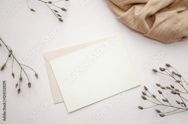 Fototapeta Blank paper card on white background with dried plants and wavy beige fabric, top view. Boho mock-up scene with greeting card template
