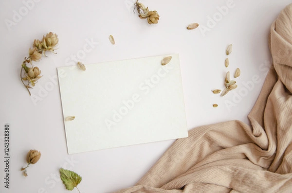 Fototapeta Blank paper card on white background with dried plants and wavy beige fabric, top view. Boho mock-up scene with greeting card template