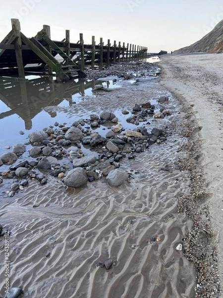 Obraz  landscape view of beautiful sandy beach with grey skies and wooden defence barrier structure pools of sea water reflect sky and chalk cliffs in background at sundown in Winter in Norfolk uk