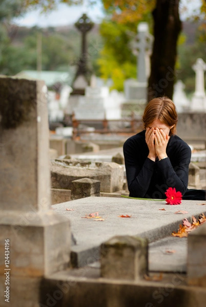 Obraz Distraught woman crying at graveside. She is covering her face with her hands. She has placed a single red flower on the stone grave.