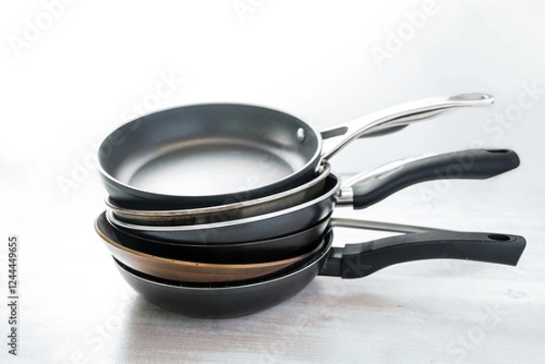 Fototapeta Stack of various frying pans made from cast iron, carbon steel, non-stick coated and stainless steel, choosing the right kitchen ware, light gray background, copy space