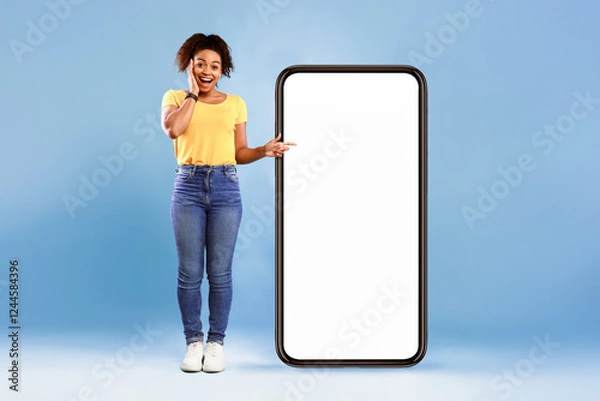 Obraz Emotional African American woman cannot believe great offer, pointing at big smartphone with space for new mobile app on screen, blue studio background. Mockup for website. Cellphone display template
