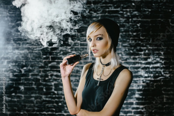 Fototapeta Sexy goth girl smokes electronic cigarette on dark background. The model vaper vaping a vaporizer in the studio.