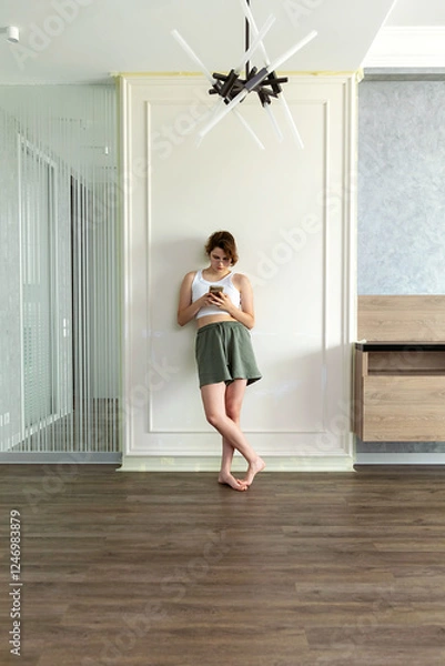 Fototapeta A teenage girl stands by an old wall ready to be painted with a cell phone. the concept of finding a professional work team to renovate an apartment.