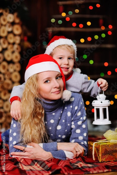 Fototapeta Mother and son in red caps of Santa Claus on a background  Christmas lights