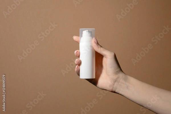 Obraz Woman's hand with manicured nails holds white bottle of cream. Unmarked container with transparent lid with pump. Background is plain, light brown in color, with slight hint of beige