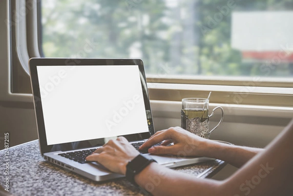 Fototapeta Close-up laptop with blank screen on table in train compartment. Women's hands on keyboard. On the table stands a cup of tea in the cup holder. In the background window. Girl using gadget. Mock up.