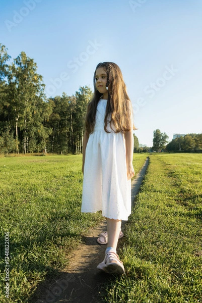 Fototapeta A young girl with long wavy brown hair stands in a sun-drenched green field in a white dress. Sunlight illuminates her hair.