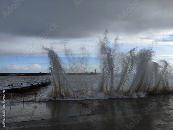 Fototapeta Waves crash against the shore creating splashes near a coastal walkway during a cloudy day