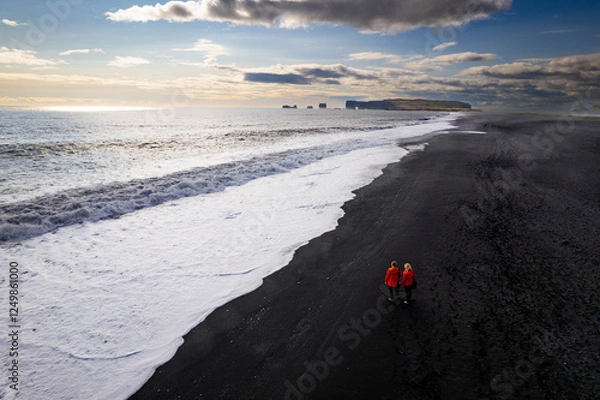 Obraz Aerail view couple walking along a stunning black sandy beach Reynisfjara , sunset in Iceland VIK Myrdal in Golden Circle area with Atlantic Ocean waves crashing and volcanic rock formations