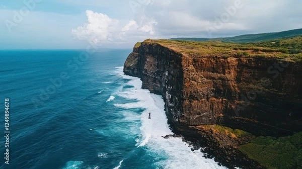 Fototapeta A stunning aerial view of a rugged cliff meeting the ocean, with waves crashing against the rock face under a cloudy sky.