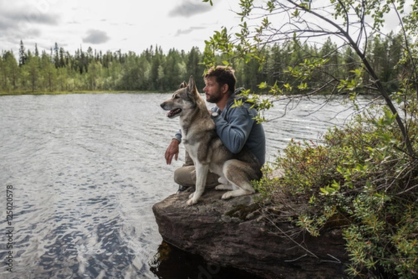 Fototapeta Handsome man and dog are sitting on the stone near of lake and looking into the distance