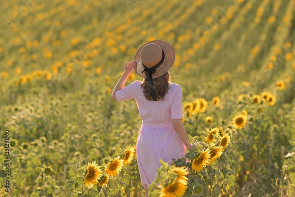 Obraz Ein Mädchen in einem Hut und einem pinken Kleid steht mit dem Rücken in einem Sonnenblumenfeld und schafft eine warme und malerische Szene.