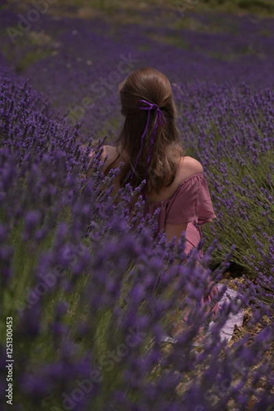 Obraz Eine Frau mit offenen Haaren und einem lila Band sitzt in einem Lavendelfeld, mit dem Rücken zur Kamera. Die Szene ist ruhig und verträumt.