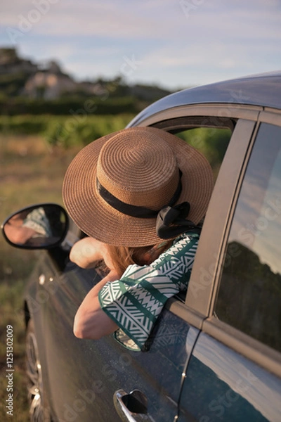 Obraz Frau in einem Hut, die aus einem Autofenster lehnt und in die Ferne blickt. Stylisches Reise- und Roadtrip-Konzept mit einem Gefühl von Freiheit und Abenteuer.