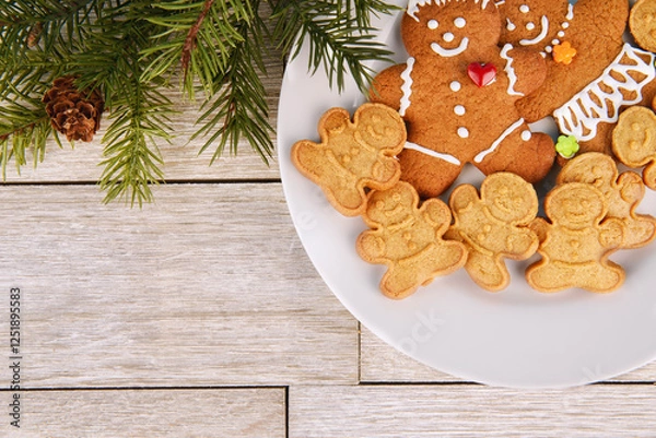 Fototapeta Festive scene featuring gingerbread cookies on a white plate, surrounded by pine branches and pinecones on a rustic wooden background. Perfect for holiday themes, ads, and cozy designs