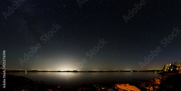 Fototapeta Panorama of the night sky above a lake in Sweden. The Milky Way is visible on the left and the bright light of large cities on the opposite side of the lake. In the foreground is the rocky coastline.