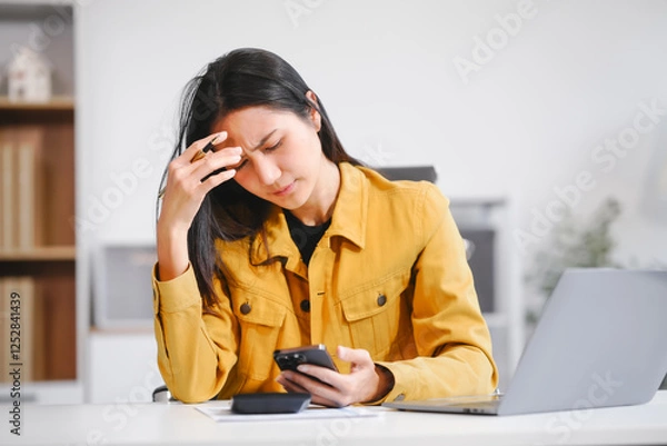 Fototapeta A businesswoman works at her desk, looking at her phone and documents. She feels stressed and experiences a migraine due to workload, deadlines, and business pressure, affecting her productivity