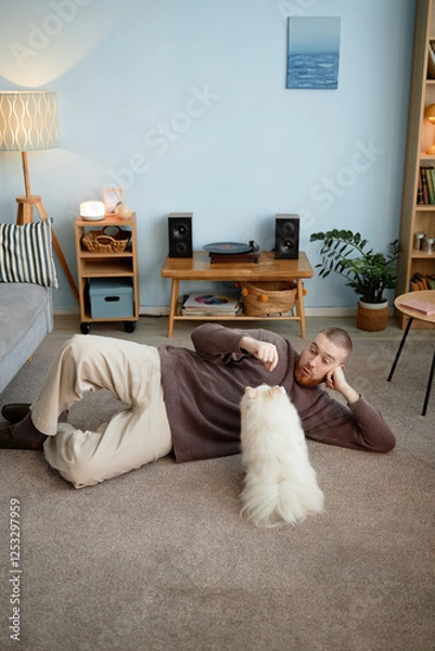 Fototapeta Person lying on carpet in living room while interacting with a fluffy dog, surrounded by cozy and stylish decor with a turntable on a wooden table and bookshelves in the background