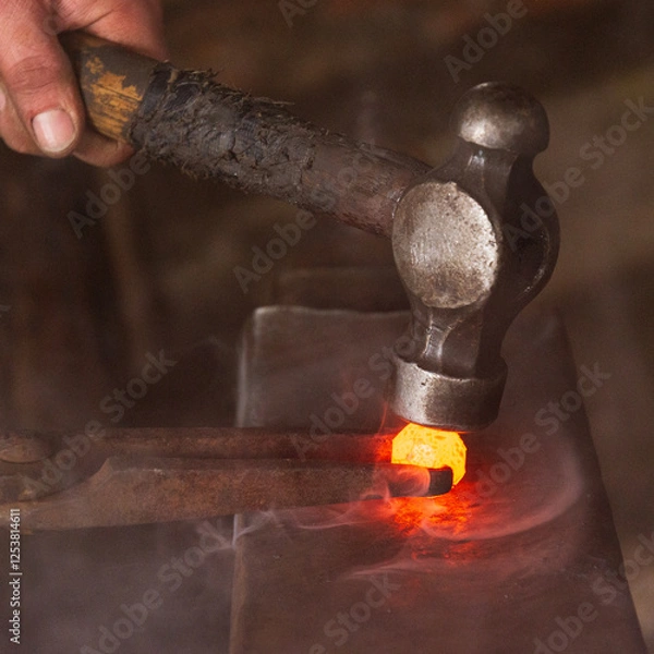 Obraz A hammer being used to shape a sphere of red hot steel held by tongs on an anvil in a forge