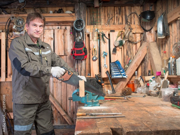 Fototapeta Adult man in work clothes and gloves cost around a wooden table with a vise and processing a piece of wood against the background of the repair shop with tools