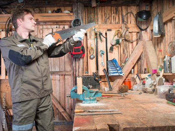 Fototapeta Adult man in work clothes and gloves cost around a wooden table with a vise and checks the blade sharpness against the background of the repair shop with tools