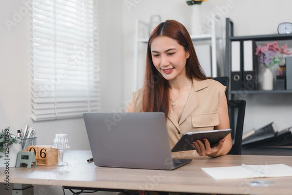 Fototapeta Woman working at desk with laptop and tablet in bright office setting.
