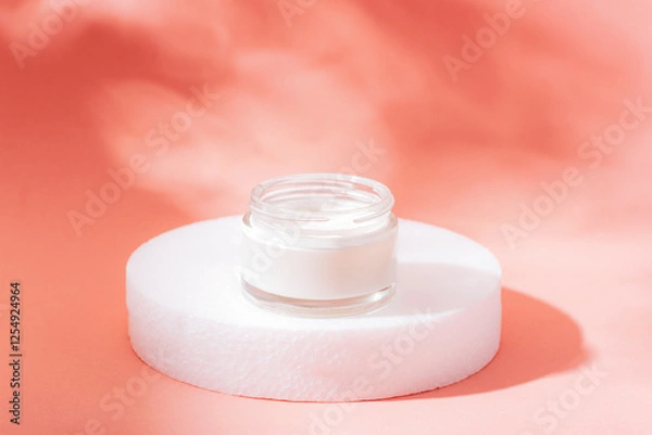 Fototapeta Glass jar of white cream on a white circular stand with a soft coral background, symbolizing skincare and beauty. Closeup